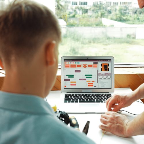 Closeup of caucasian teacher hand picking wires and teaching about electronic equipment while boy looking at instructor using electric tool at table with laptop display coding screen. Edification.